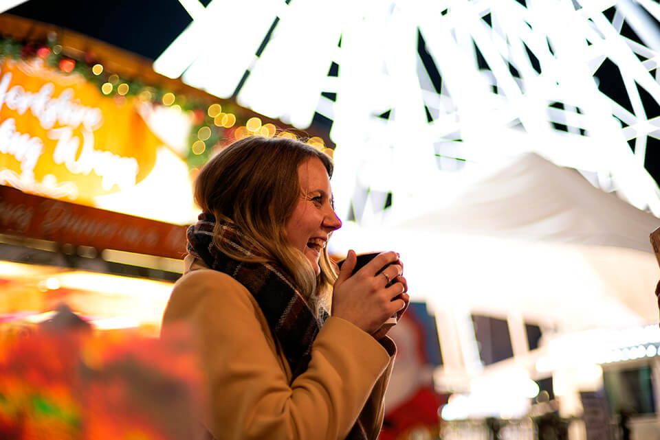 A woman wearing a scarf laughing and enjoying a hot drink under the Wheel of Light at Jubilee Square in Leicester