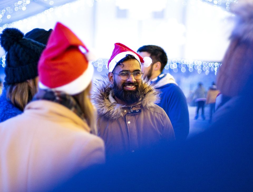 A smiling man with a beard among a group wearing christmas hats while ice skating