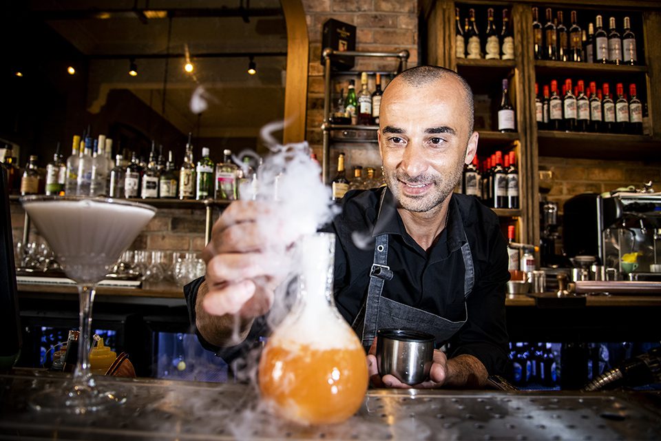 A man behind a bar making a cocktail in a decorative glass with smoke pouring out