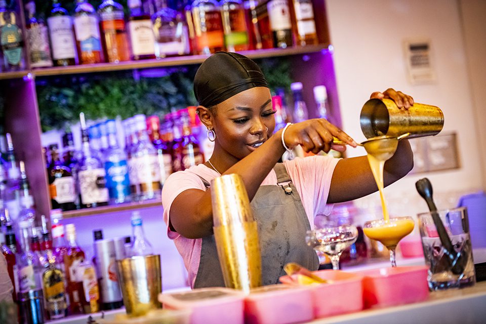 A woman behind a bar pouring a drink from a shaker into a cocktail glass